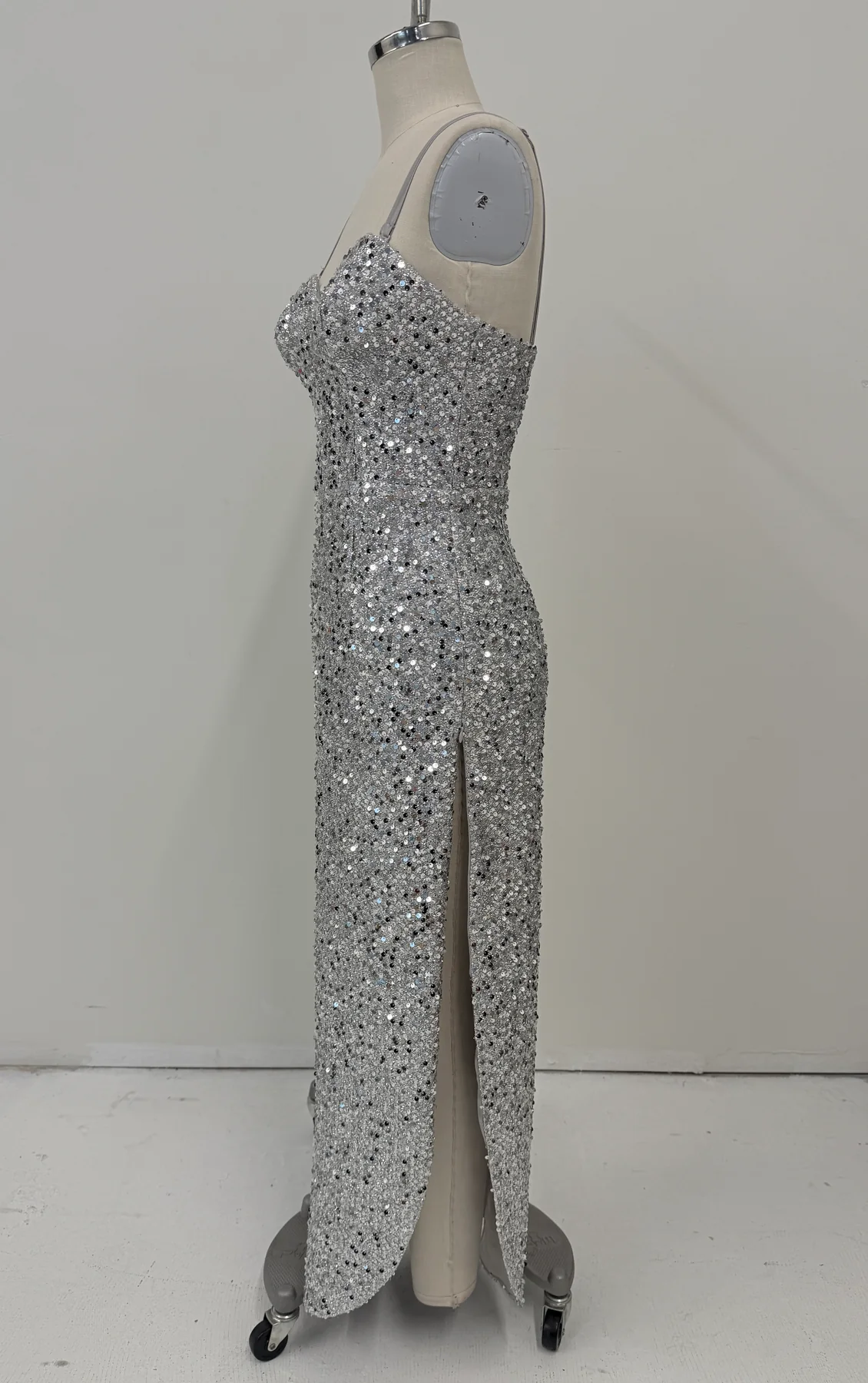 A floor-length silver sequined gown displayed on a beige mannequin dress form against a plain light gray wall and floor.