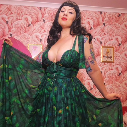 A woman modeling a dark green floor-length dress with a leaf print, holding out the sheer sides of the skirt against a pink flamingo-patterned wallpaper background.