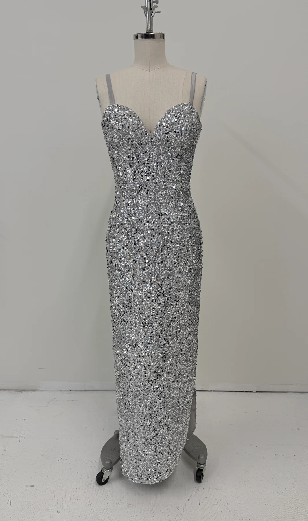 A floor-length silver sequin gown displayed on a dress form mannequin against a plain light gray wall and white floor.