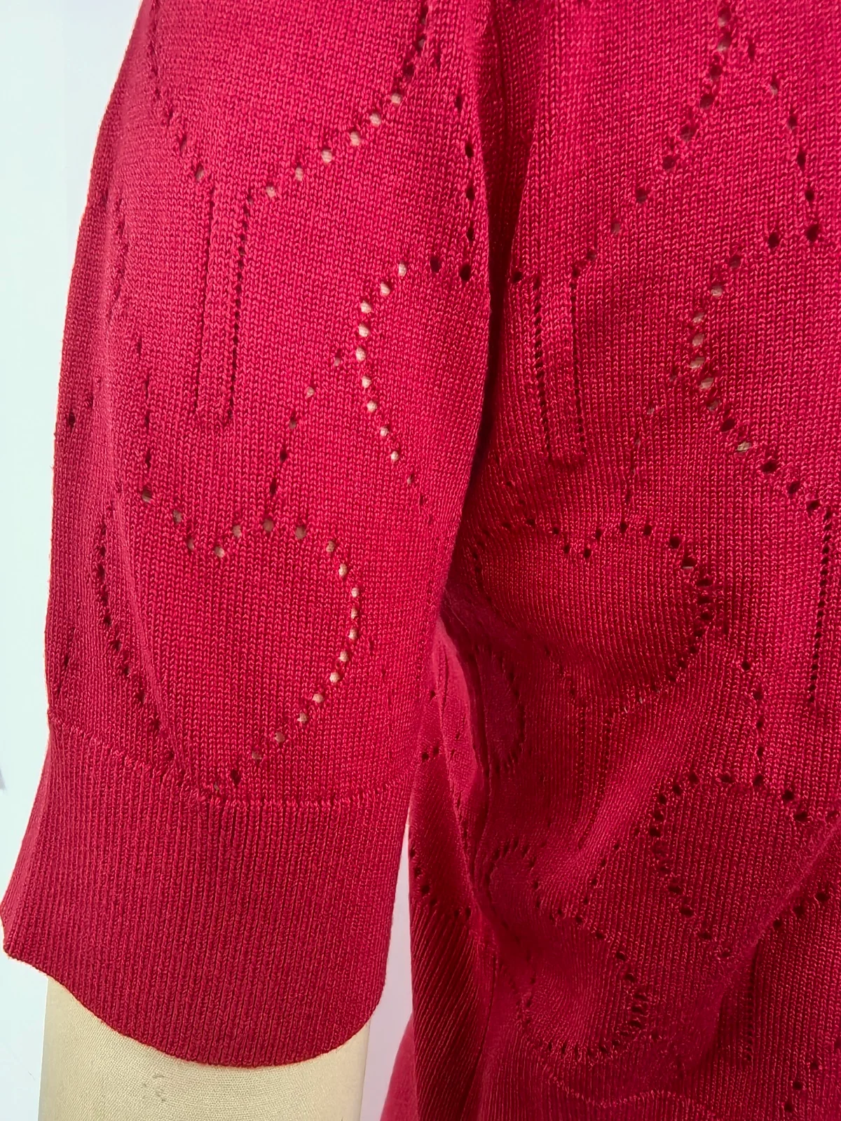 A close-up of a bright red knit top or sweater sleeve with an eyelet heart pattern and a ribbed cuff, shown on a mannequin arm against a plain light background.