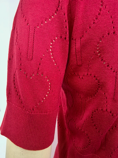 A close-up of a bright red knit top or sweater sleeve with an eyelet heart pattern and a ribbed cuff, shown on a mannequin arm against a plain light background.