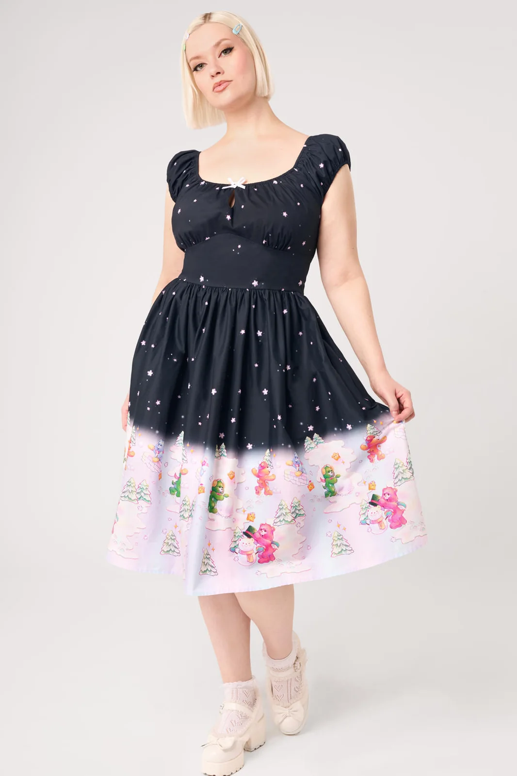 A woman modeling a short black dress with white star-like speckles and a pastel winter scene printed along the hem, paired with white lace-up shoes, against a light gray studio background.