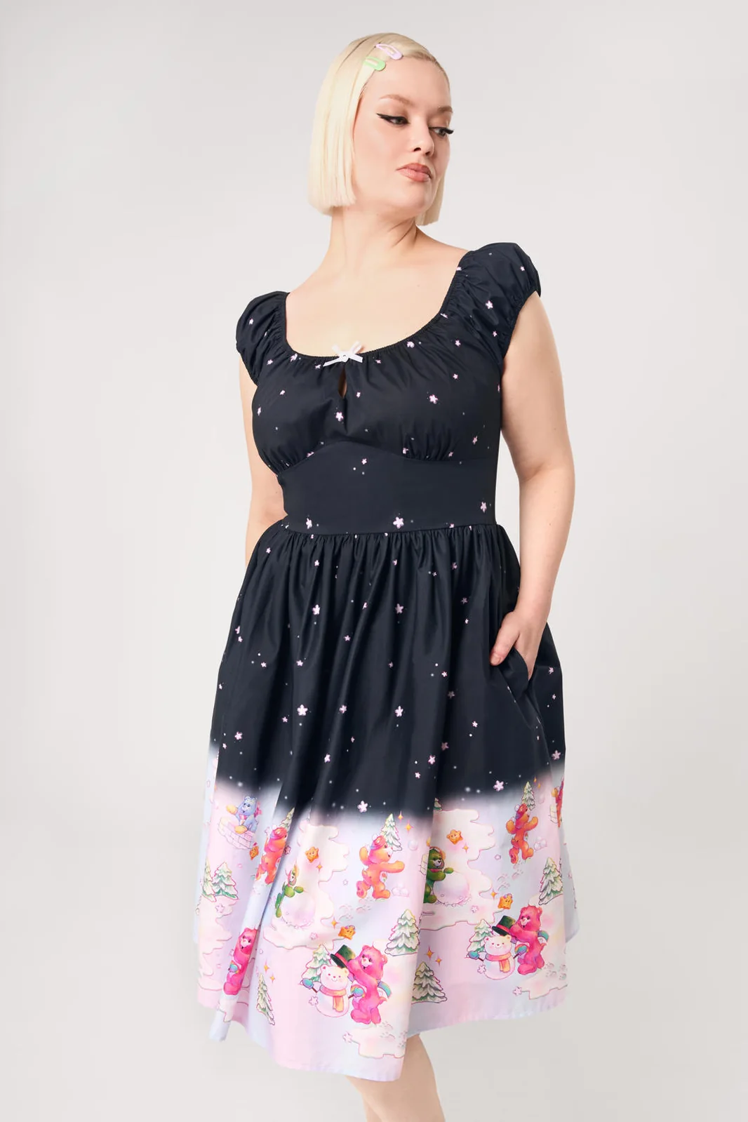 A woman modeling a short-sleeve midi dress with a black upper section featuring a white star pattern, and a light pink lower section with a colorful winter print of snowmen, Santa Claus, and snow globes.
