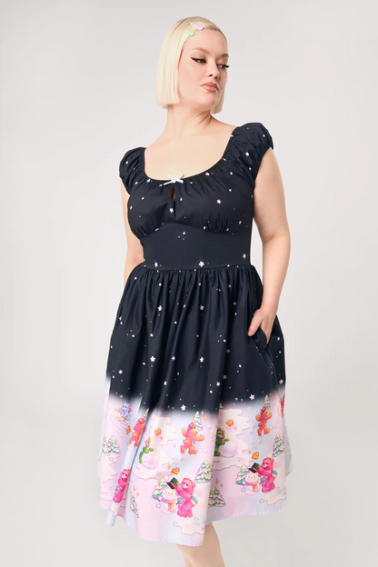 A woman modeling a short-sleeve midi dress with a black upper section featuring a white star pattern, and a light pink lower section with a colorful winter print of snowmen, Santa Claus, and snow globes.