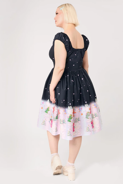 A woman modeling a short black dress with white star-like speckles and a pastel gradient hem featuring a whimsical winter print, shown from the back against a light gray studio background.