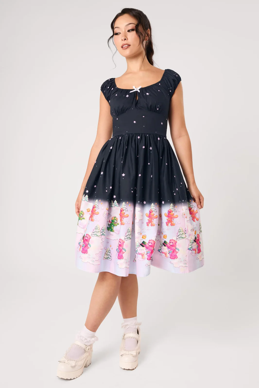 A woman modeling a navy blue A-line dress with a white star pattern on the upper half and a light pink lower half featuring colorful gingerbread men illustrations, paired with white lace-up shoes.