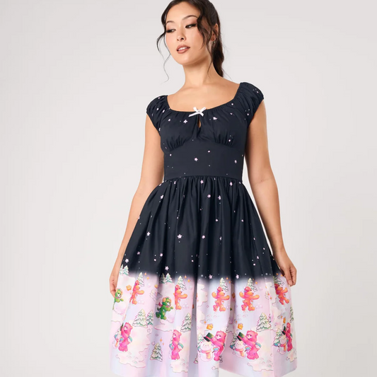 A woman modeling a short-sleeve midi dress with a navy bodice printed with white stars, fading into a light pink skirt with a gingerbread man and snowman winter print.