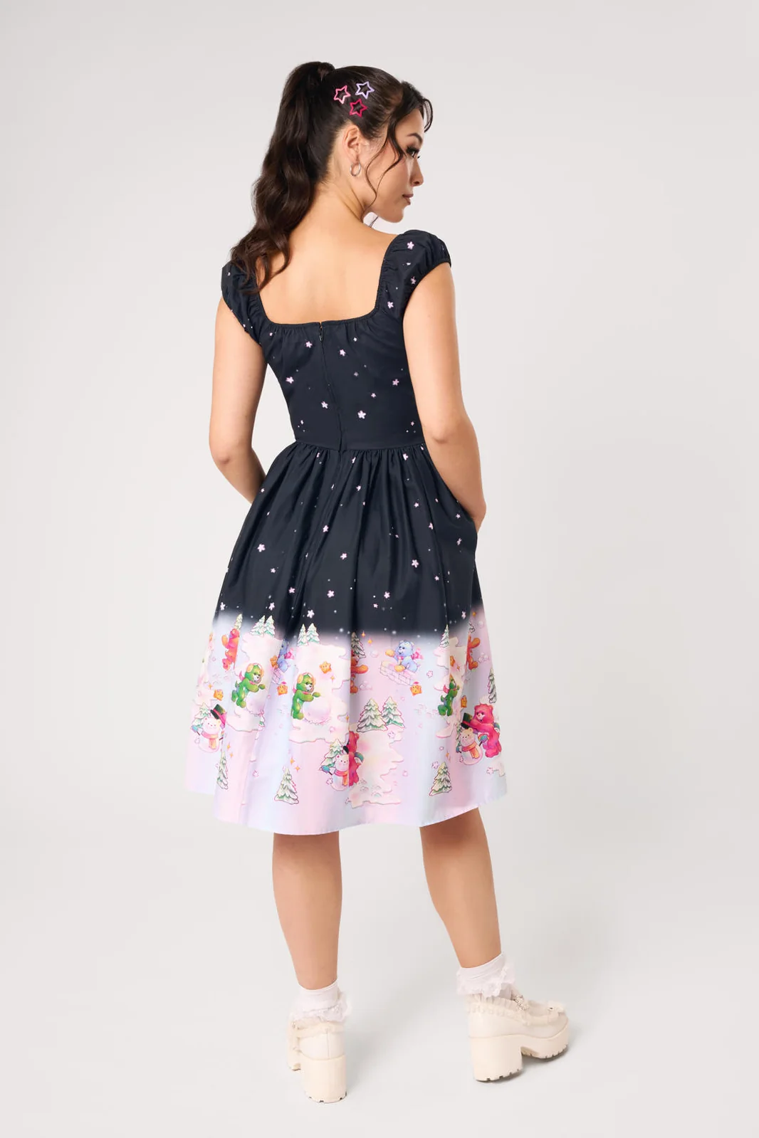 A woman modeling a short dress from the back against a light gray studio background. The dress has a dark navy upper section with small white star-like dots, and a light pink lower section with a colorful floral and whimsical print.