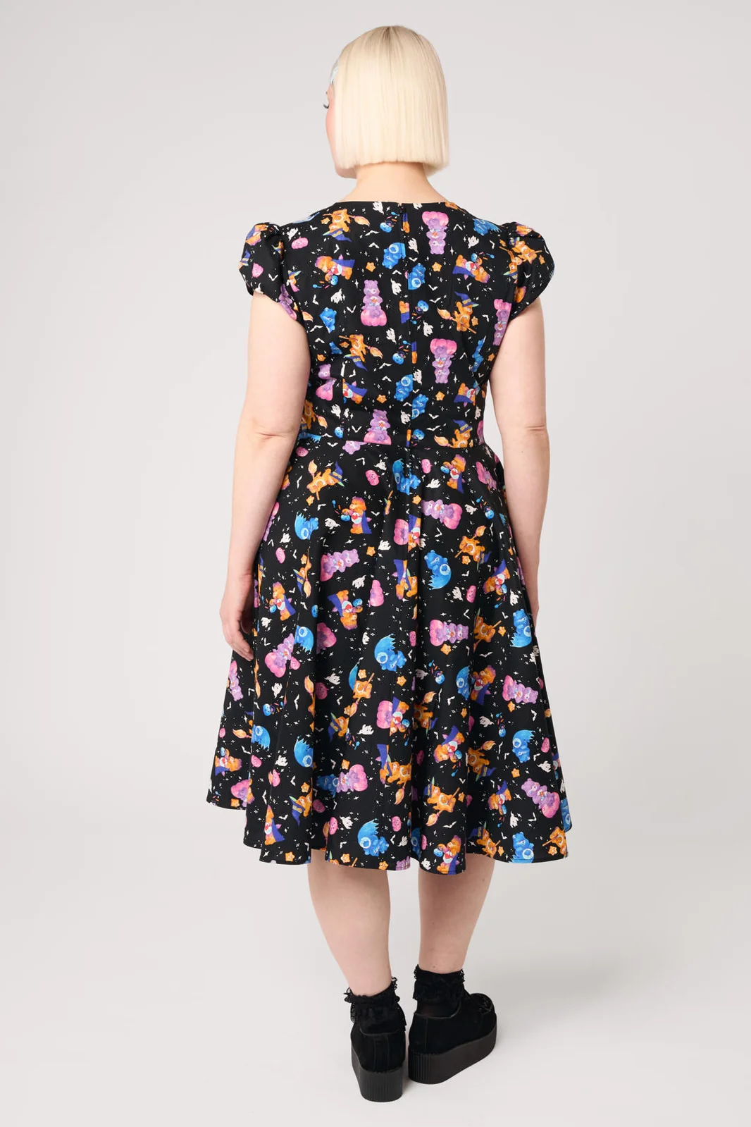 A woman viewed from the back wearing a black midi dress with a colorful cartoon character print, puff sleeves, and black platform shoes against a light gray studio background.