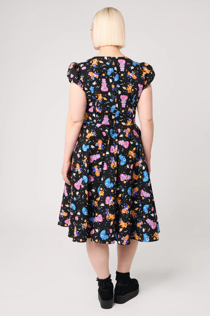 A woman viewed from the back wearing a black midi dress with a colorful cartoon character print, puff sleeves, and black platform shoes against a light gray studio background.