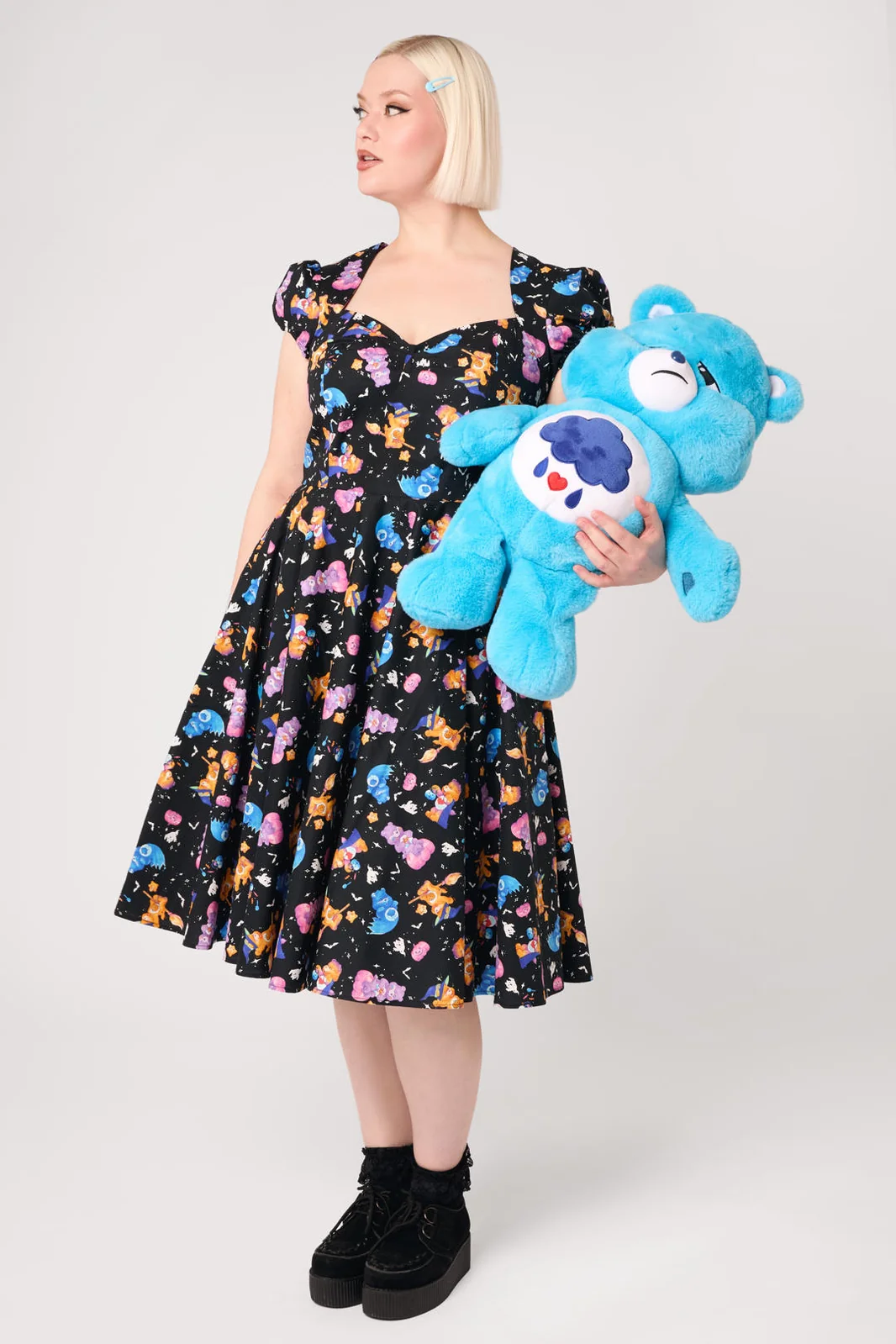 A woman modeling a black scoop-neck swing dress with a colorful cartoon bear print, holding a blue Care Bear plush toy against a light gray studio background.