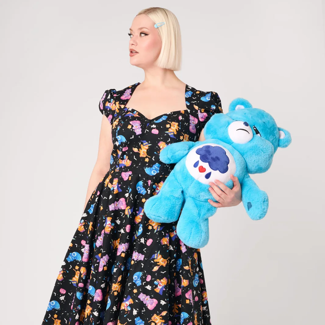 A blonde woman wearing a black dress with a colorful cartoon bear print holds a large blue plush teddy bear with a white belly patch on a light gray background.