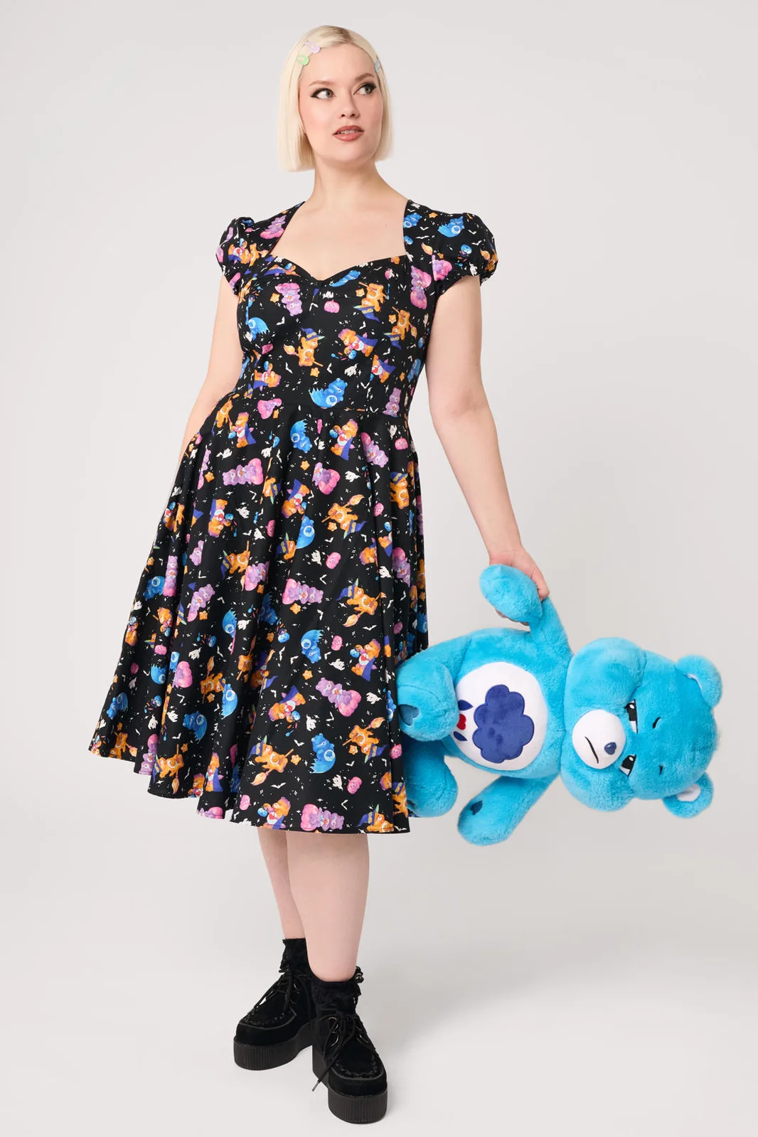 A woman modeling a black midi dress with a colorful cartoon character print, featuring puff sleeves, a sweetheart neckline, and a flared skirt, paired with white socks and black Mary Jane shoes against a light gray studio background.