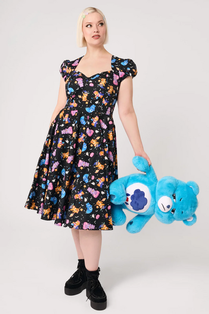 A woman modeling a black midi dress with a colorful cartoon character print, featuring puff sleeves, a sweetheart neckline, and a flared skirt, paired with white socks and black Mary Jane shoes against a light gray studio background.
