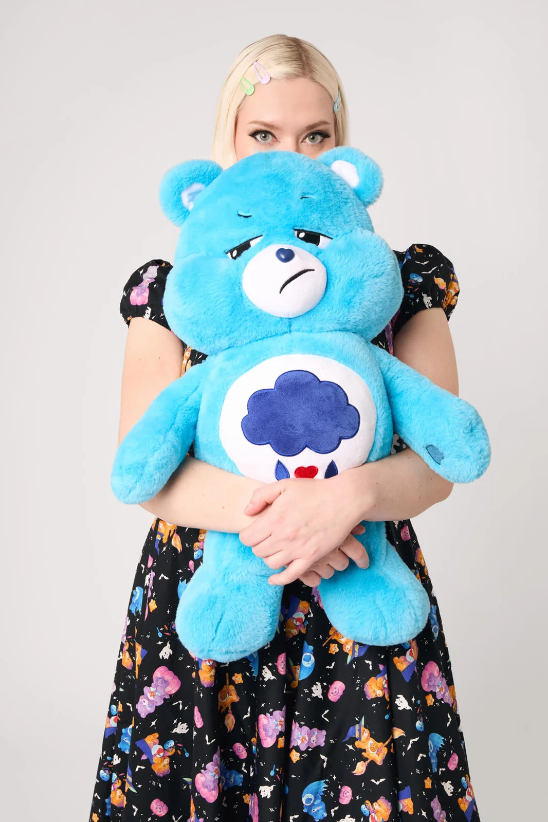 A blonde woman wearing a black floral dress holds a large blue plush bear with a sad face and a dark blue cloud emblem on its chest, set against a plain light gray background.