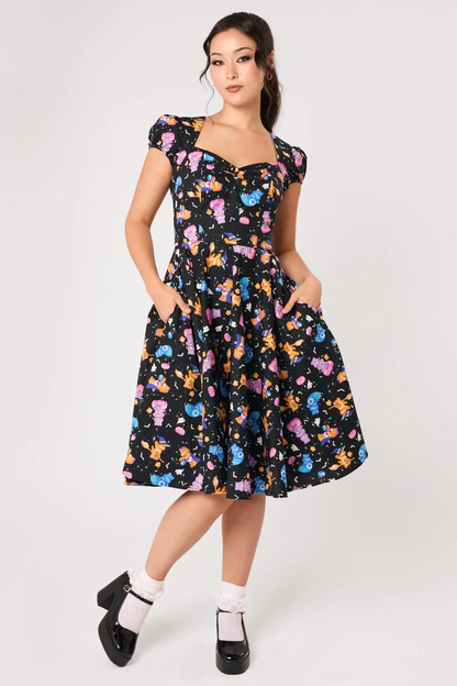 A woman modeling a black midi dress with a colorful cartoon character print, featuring puff sleeves, a sweetheart neckline, and a flared skirt, paired with white socks and black Mary Jane shoes against a light gray studio background.