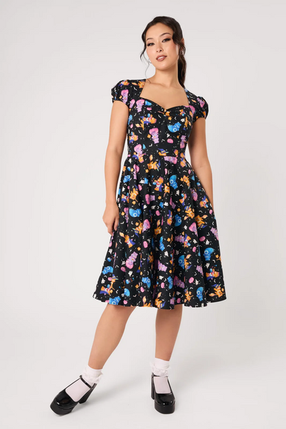 A woman modeling a black midi dress with a colorful cartoon character print, featuring puff sleeves, a sweetheart neckline, and a flared skirt, paired with white socks and black Mary Jane shoes against a light gray studio background.