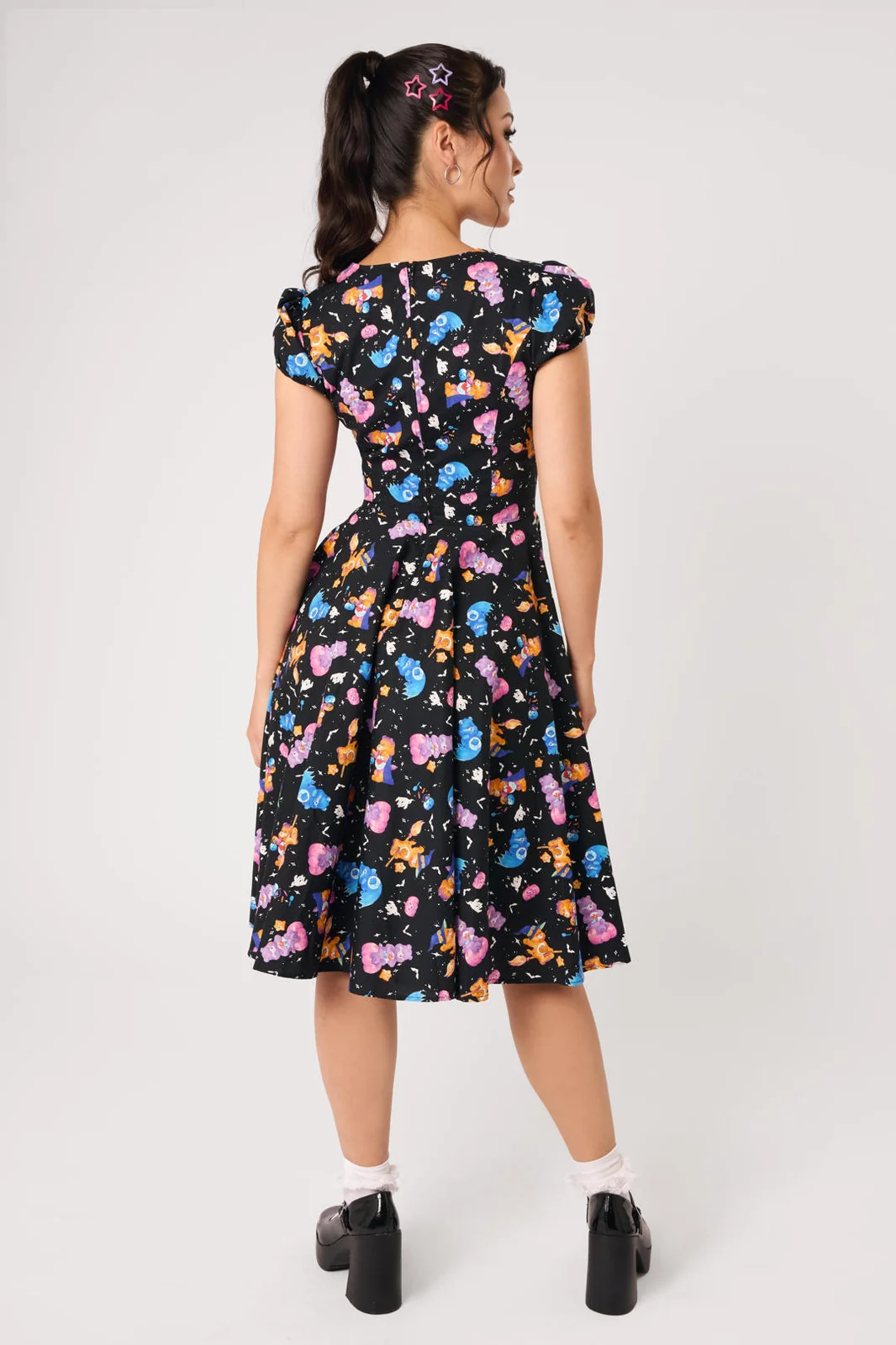 A woman modeling a black midi dress with a colorful cartoon character print, featuring puff sleeves, a sweetheart neckline, and a flared skirt, paired with white socks and black Mary Jane shoes against a light gray studio background.
