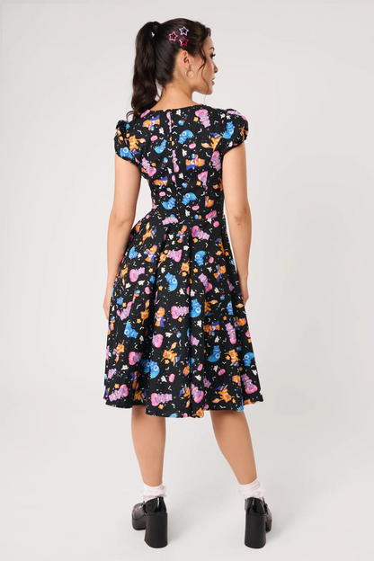 A woman modeling a black midi dress with a colorful cartoon character print, featuring puff sleeves, a sweetheart neckline, and a flared skirt, paired with white socks and black Mary Jane shoes against a light gray studio background.
