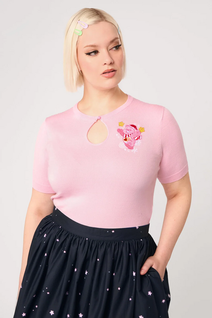 A blonde model wearing a light pink short-sleeve knit top with a keyhole cutout and a Piglet embroidery on the chest, paired with a dark navy skirt with small white star prints, against a light gray studio background.