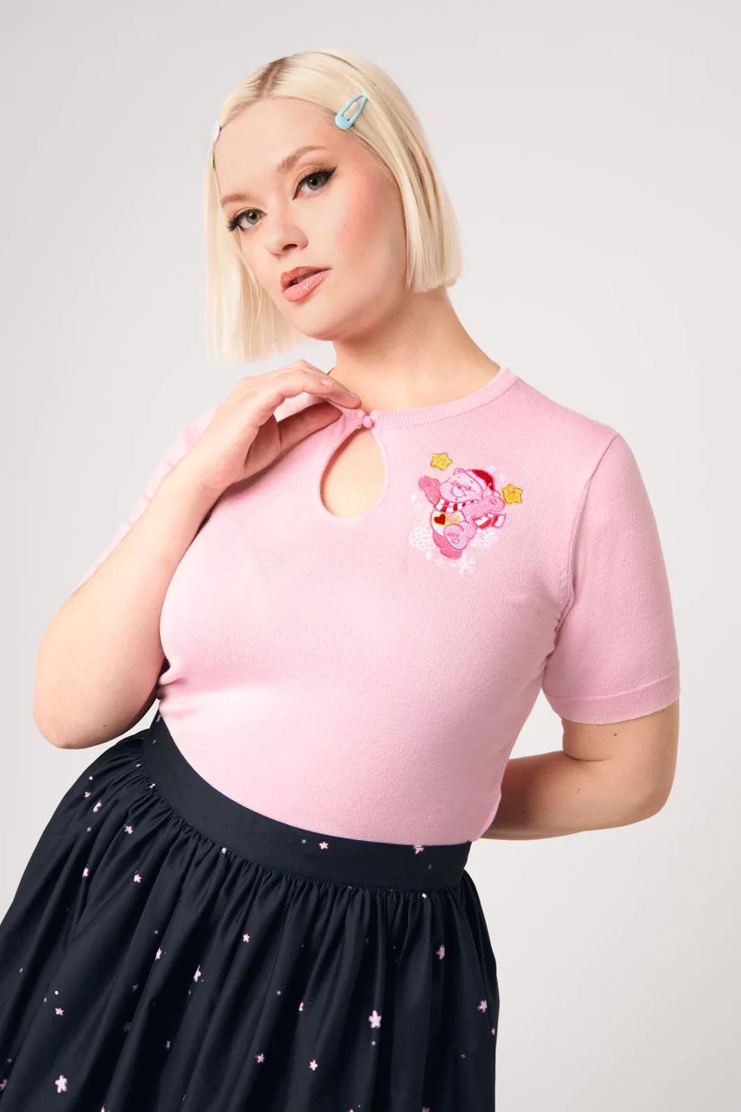 A blonde model wearing a light pink short-sleeve knit top with a keyhole cutout and a pink cartoon character graphic on the chest, paired with a dark navy skirt with small star prints, against a light gray studio background.