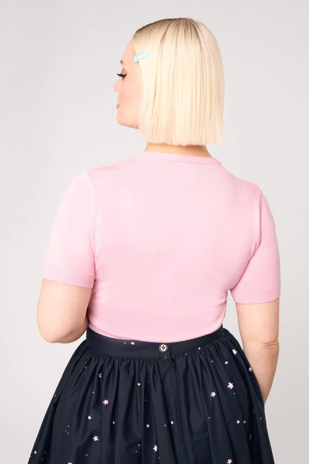 A woman with a short blonde bob is shown from the back, wearing a light pink short-sleeve knit top and a black skirt with small pink star prints, against a light gray studio background.