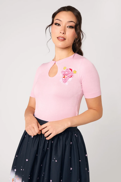 A woman modeling a light pink short-sleeve top with a keyhole cutout and a pink cartoon graphic on the chest, paired with a dark navy skirt with small star prints, against a light gray studio background.