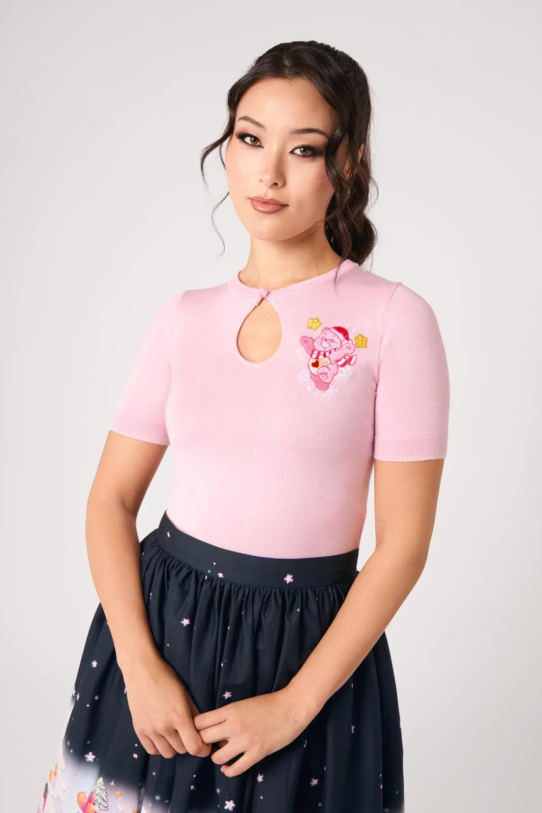 A woman modeling a light pink short-sleeve top with a keyhole neckline and a pink cartoon character graphic on the left chest, paired with a dark navy skirt with a star pattern, against a light gray studio background.