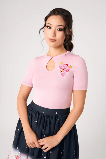 A woman modeling a light pink short-sleeve top with a keyhole neckline and a pink cartoon character graphic on the left chest, paired with a dark navy skirt with a star pattern, against a light gray studio background.