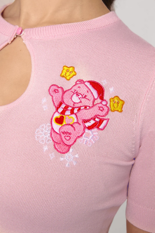A close-up of a pink knit top featuring a large embroidered pink bear character wearing a Santa hat and scarf, with yellow star accents, on the chest.