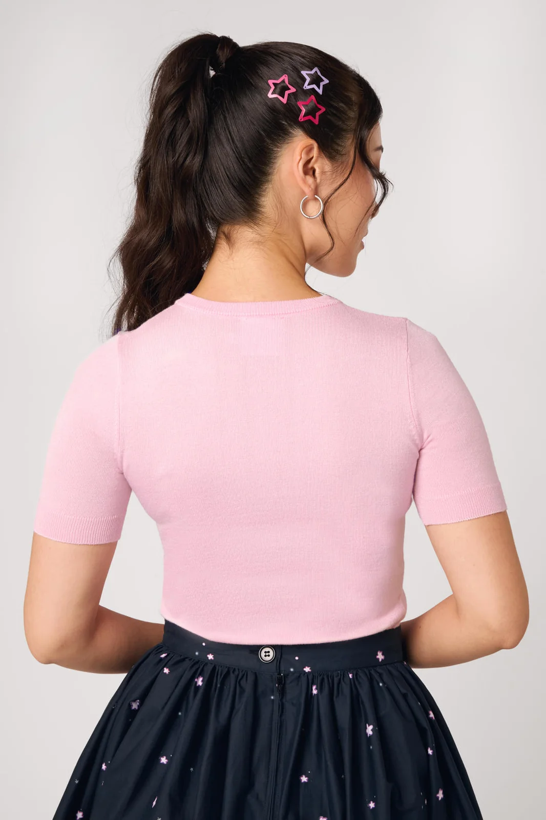 A woman shown from the back wearing a light pink short-sleeve crewneck top and a dark navy skirt with small pink star prints. She has her hair in a high ponytail with pink and silver star hair clips.