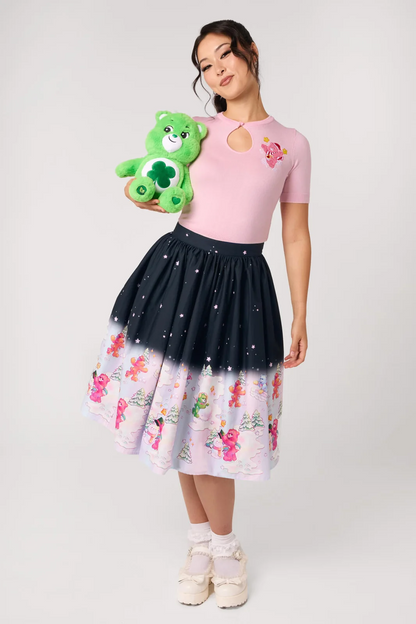 A woman modeling a pink short-sleeve top and a dark navy-to-pale pink ombré skirt with Care Bear illustrations, holding a green plush Care Bear toy against a light gray studio background.