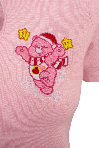 A close-up of a light pink ribbed knit garment featuring an embroidered pink bear character wearing a Santa hat and scarf, with yellow star accents.