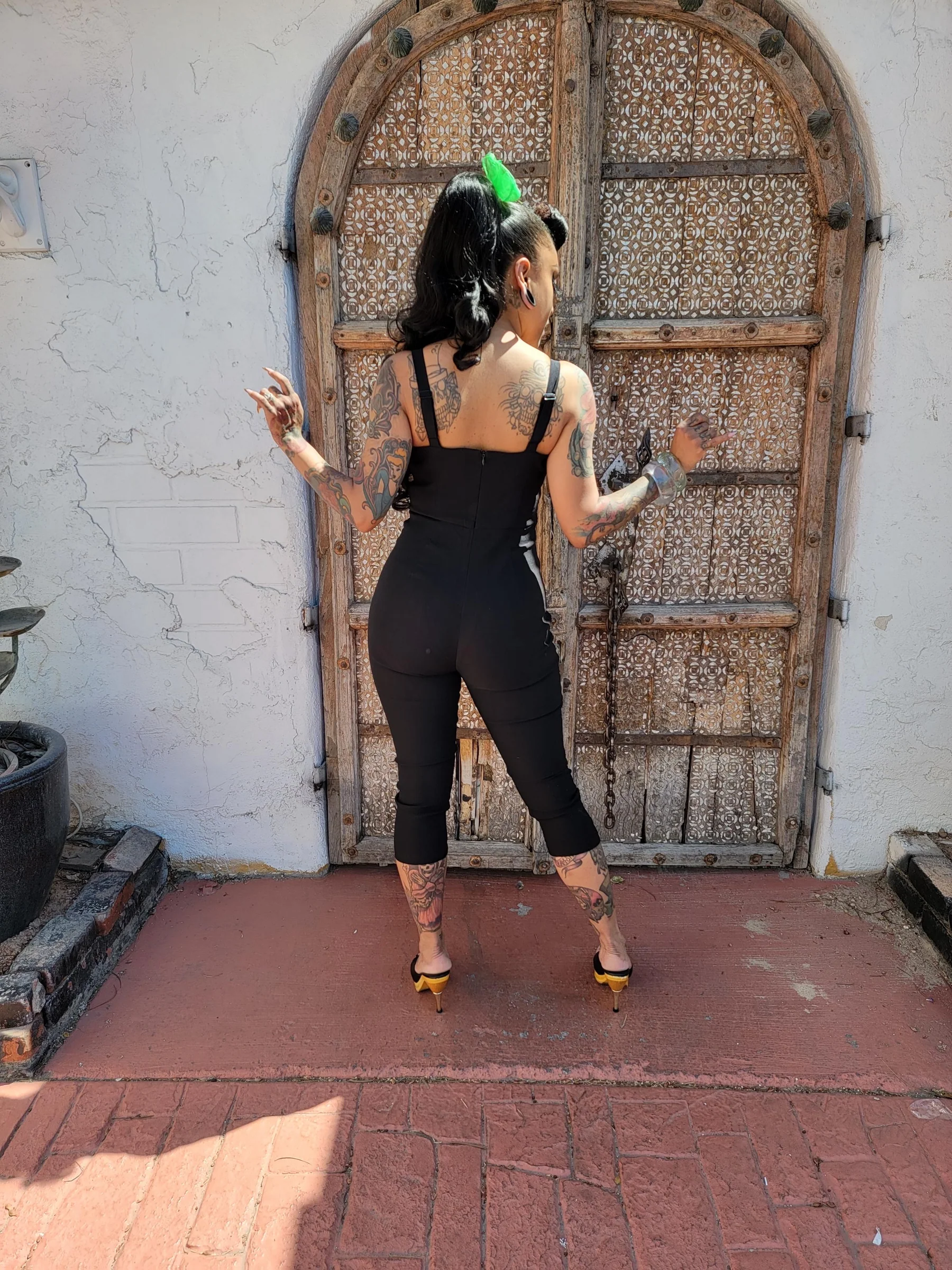 A woman stands with her back to the camera in front of a large rustic wooden door, wearing a black sleeveless jumpsuit and yellow high heels.