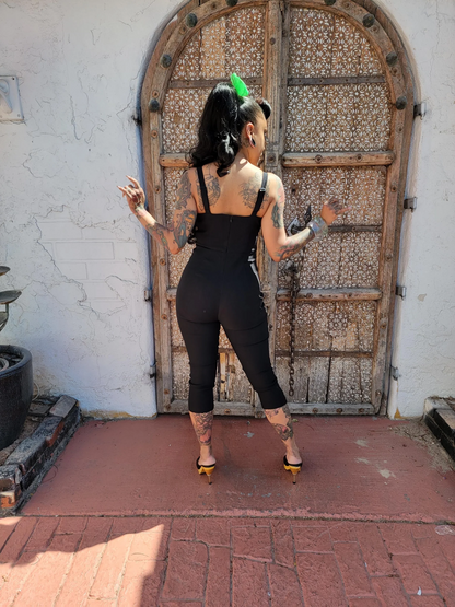 A woman stands with her back to the camera in front of a large rustic wooden door, wearing a black sleeveless jumpsuit and yellow high heels.