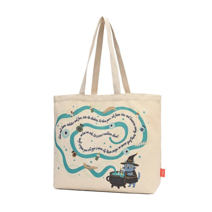 A beige canvas tote bag featuring a whimsical printed design of a winding teal path with text, stars, and a small blue cat in a witch hat next to a cauldron.