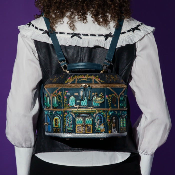 A person viewed from the back wearing a decorative illustrated backpack with a dark teal and black base, gold trim, and colorful architectural building motifs. The person wears a white ruffled blouse and black vest against a solid purple background.