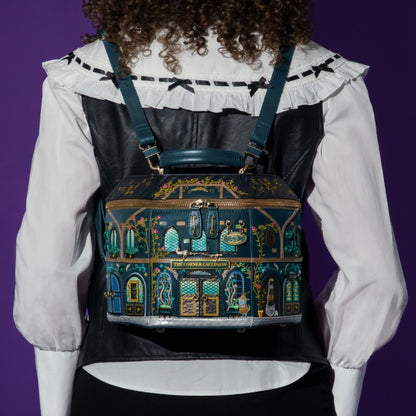 A person viewed from the back wearing a decorative illustrated backpack with a dark teal and black base, gold trim, and colorful architectural building motifs. The person wears a white ruffled blouse and black vest against a solid purple background.