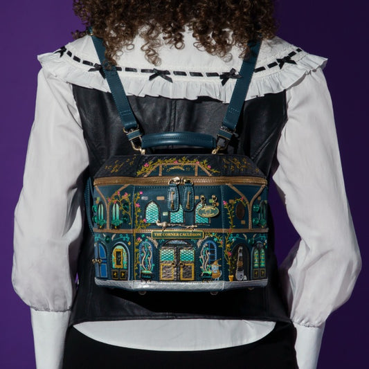A person viewed from the back wearing a decorative illustrated backpack with a dark teal and black base, gold trim, and colorful architectural building motifs. The person wears a white ruffled blouse and black vest against a solid purple background.