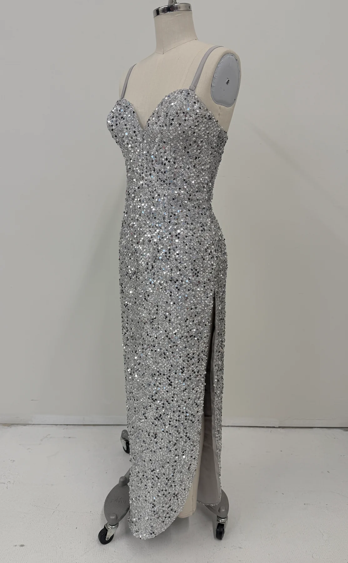 A floor-length silver sequin gown displayed on a mannequin with a high slit, photographed against a plain light gray wall and floor.