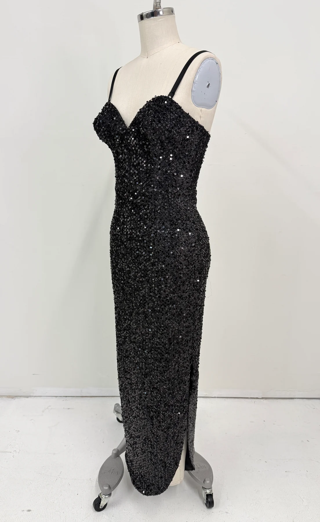 A black sequined evening gown displayed on a dress form mannequin. The sleeveless gown features thin spaghetti straps, a sweetheart neckline, a fitted silhouette, and a high thigh slit.