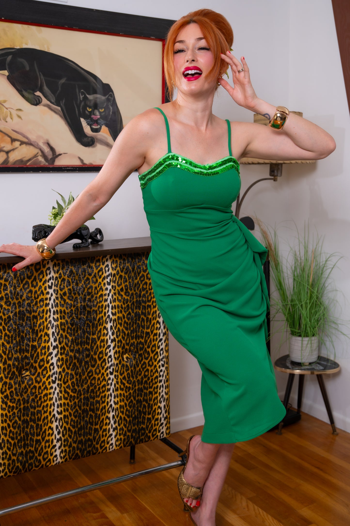 A woman with red hair wearing a bright green spaghetti-strap dress with sequined trim, posing in a room with a leopard-print cabinet, a framed panther painting, and wooden floors.