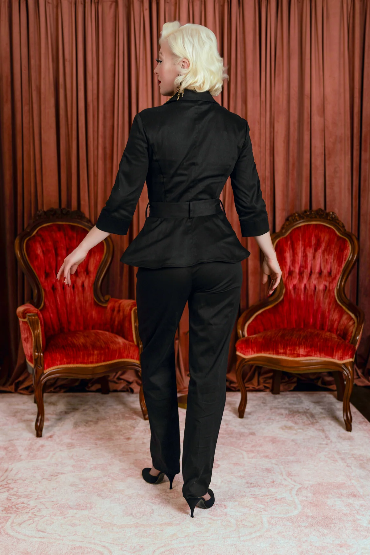 A woman with short blonde hair is shown from behind wearing a black tailored pantsuit with a peplum waist, three-quarter sleeves, and a matching belt. She is standing in a room with red velvet armchairs and a pink patterned rug in front of a rust-colored curtain.