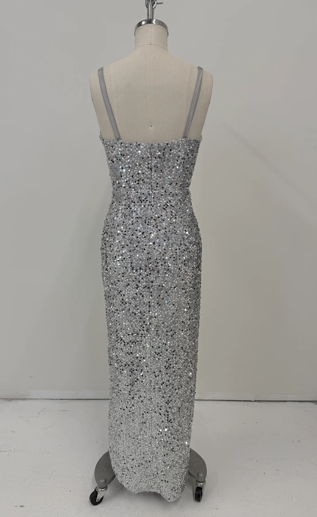 A floor-length silver sequined gown displayed on a dress form mannequin against a plain light gray studio wall and floor.