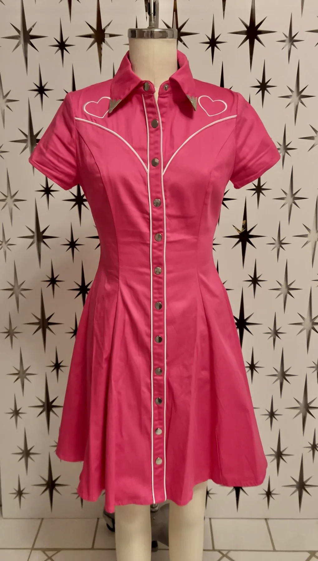 Pink dress with heart designs on a mannequin against a patterned wall.