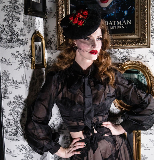 Woman in a dark, Victorian-style outfit with red flowers against a patterned wall.
