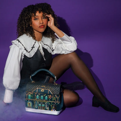 A model wearing a white ruffled blouse and a black pinafore dress sits on a purple studio floor, next to a decorative handbag featuring a colorful illustrated house design.
