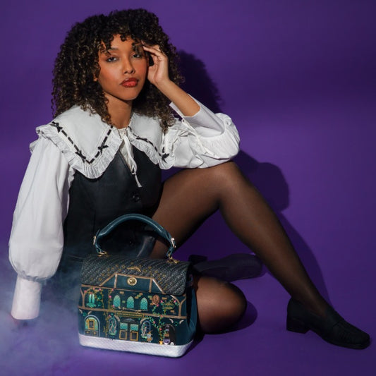 A model wearing a white ruffled blouse and a black pinafore dress sits on a purple studio floor, next to a decorative handbag featuring a colorful illustrated house design.