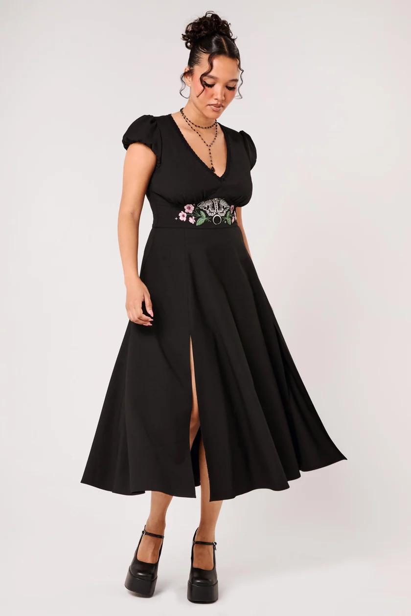 A woman modeling a black midi dress with short puff sleeves, a V-neckline, and a high thigh slit. The dress features a decorative embroidered waistband with pink, white, and green floral details.