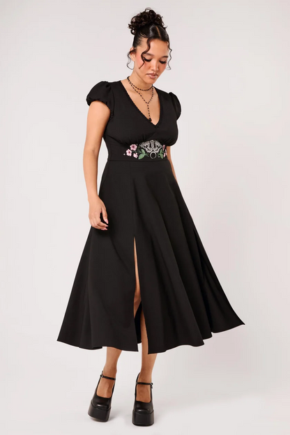 A woman modeling a black midi dress with short puff sleeves, a V-neckline, and a high thigh slit. The dress features a decorative embroidered waistband with pink, white, and green floral details.